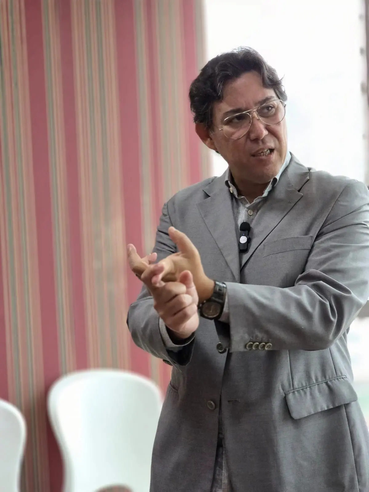 Fabio Blanco gesticulando com as duas mãos enquanto explica, de paletó e microfone de lapela